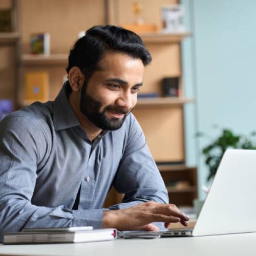 Smiling indian business man working on laptop at home office. Young indian student or remote teacher using computer remote studying, virtual training, watching online education webinar at home office.